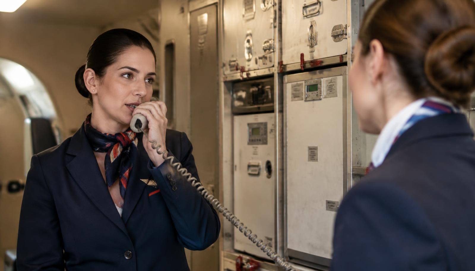 A cabin crew member speaking on the interphone in an aircraft galley while a colleague listens
